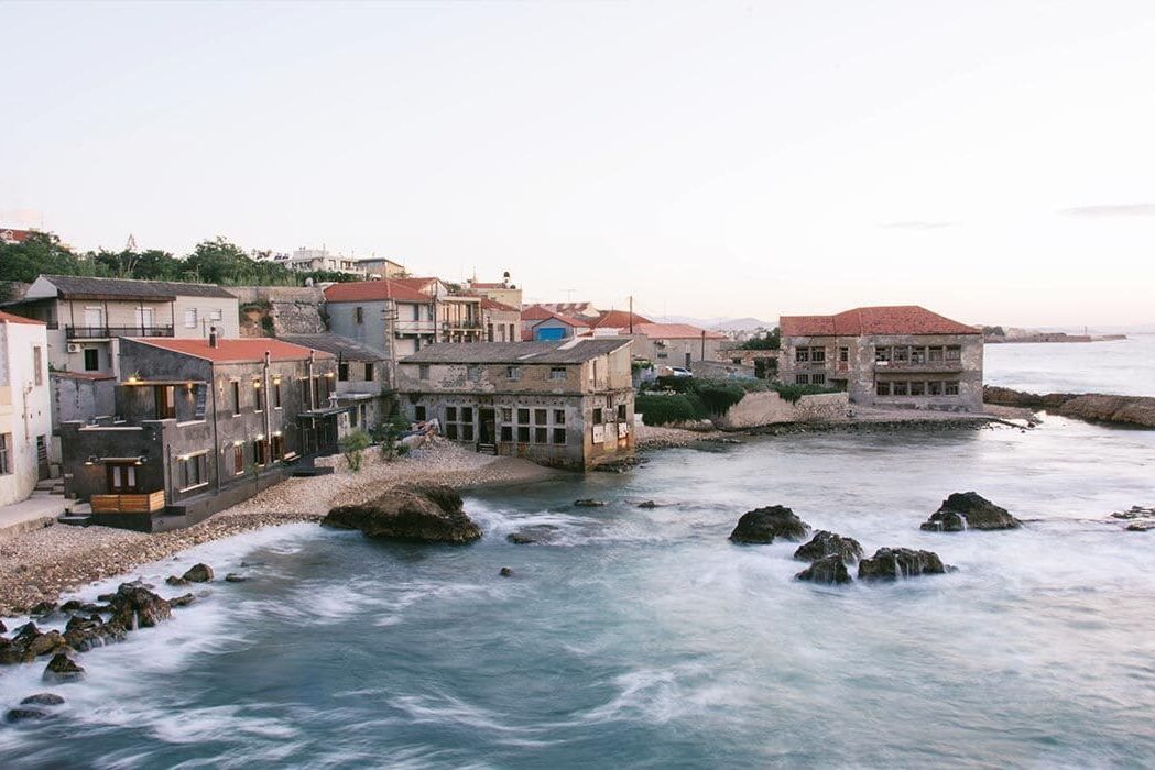 The row of tanneries seen from the water at dusk, waves breaking over the rocks in front of them