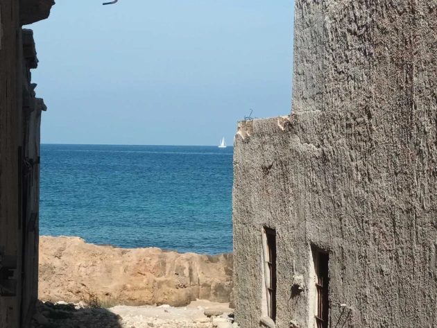 A narrow gap between two tannery walls opening onto blue sea, a sailing boat on the horizon