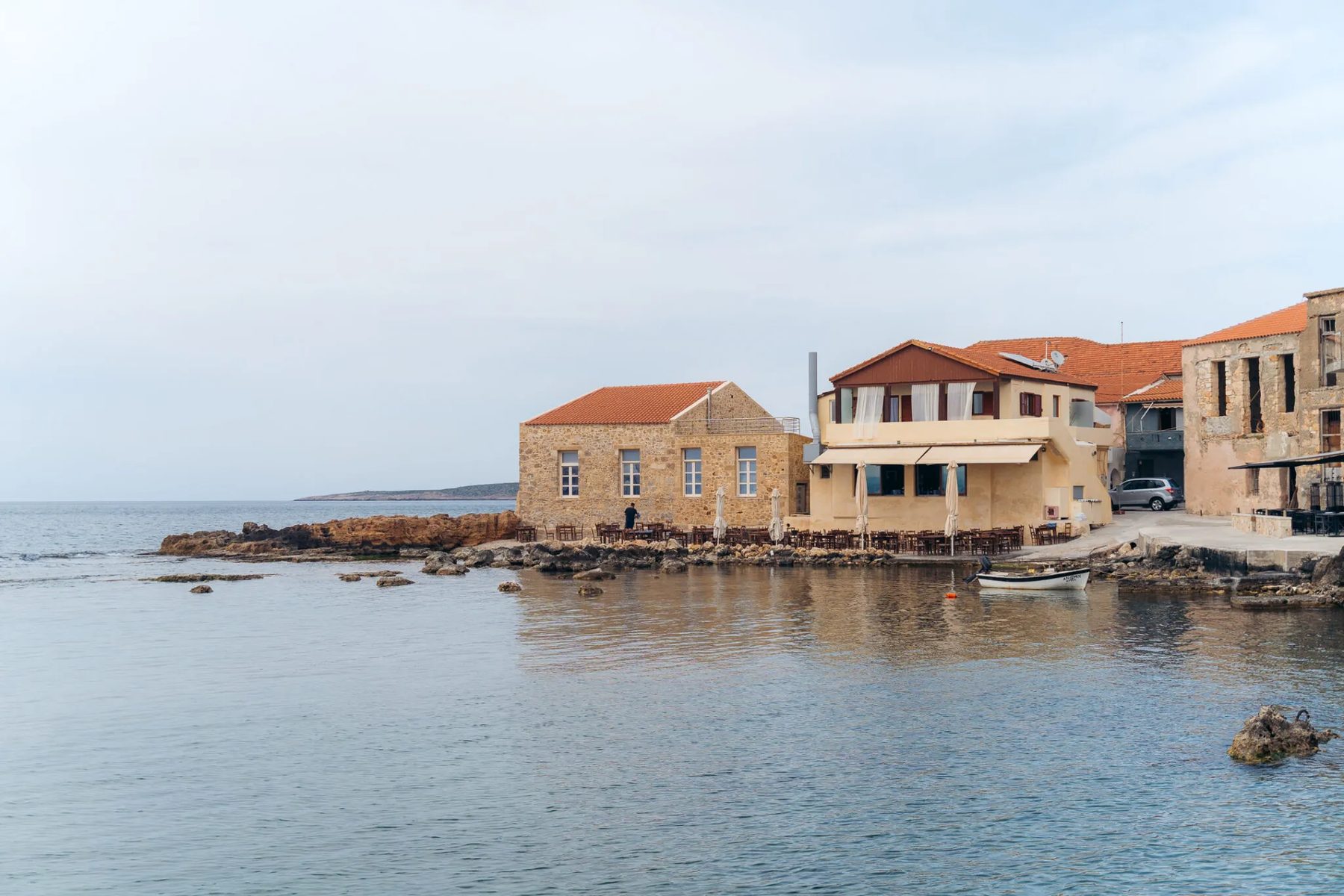 Restored stone buildings on the water with tables set out on the rocks and a small boat moored alongside