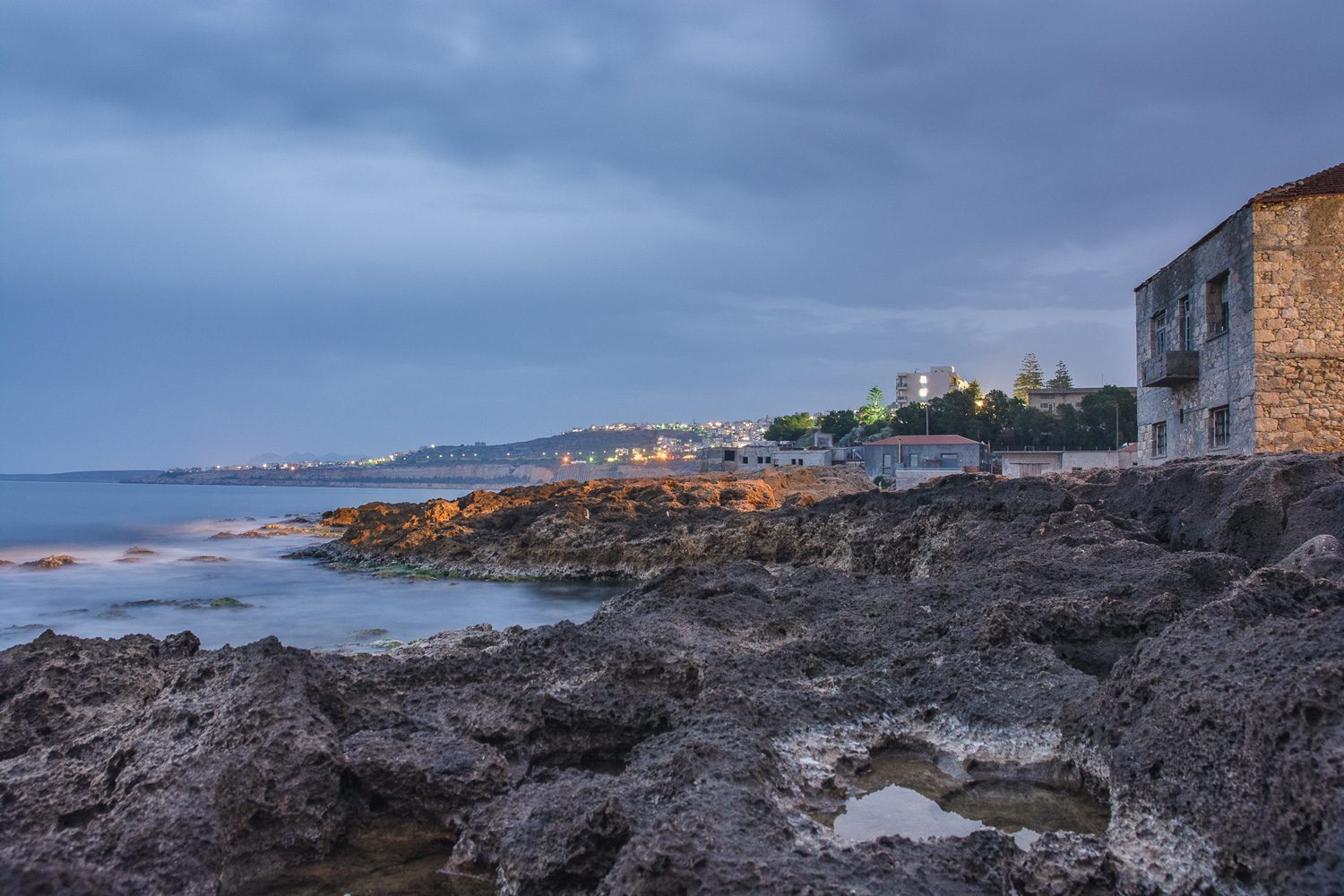 The rocky shore at dusk, water smoothed by a long exposure, the lights of the town along the coast